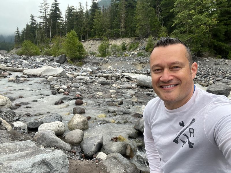 Smiling man in white shirt near a rocky river in a forested area.